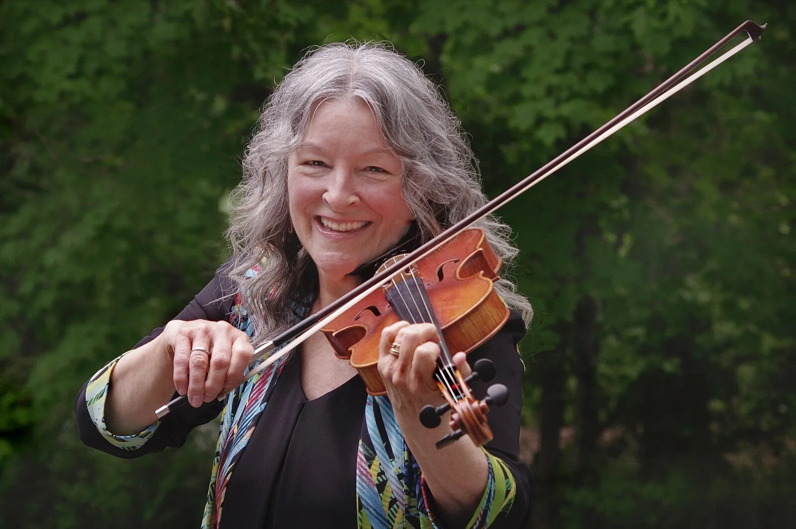 A photo of Cindy Thompson smiling and playing her fiddle outdoors.