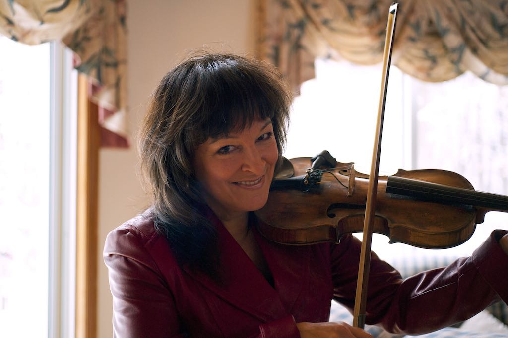 Cindy smiling and playing the fiddle in front of two windows.