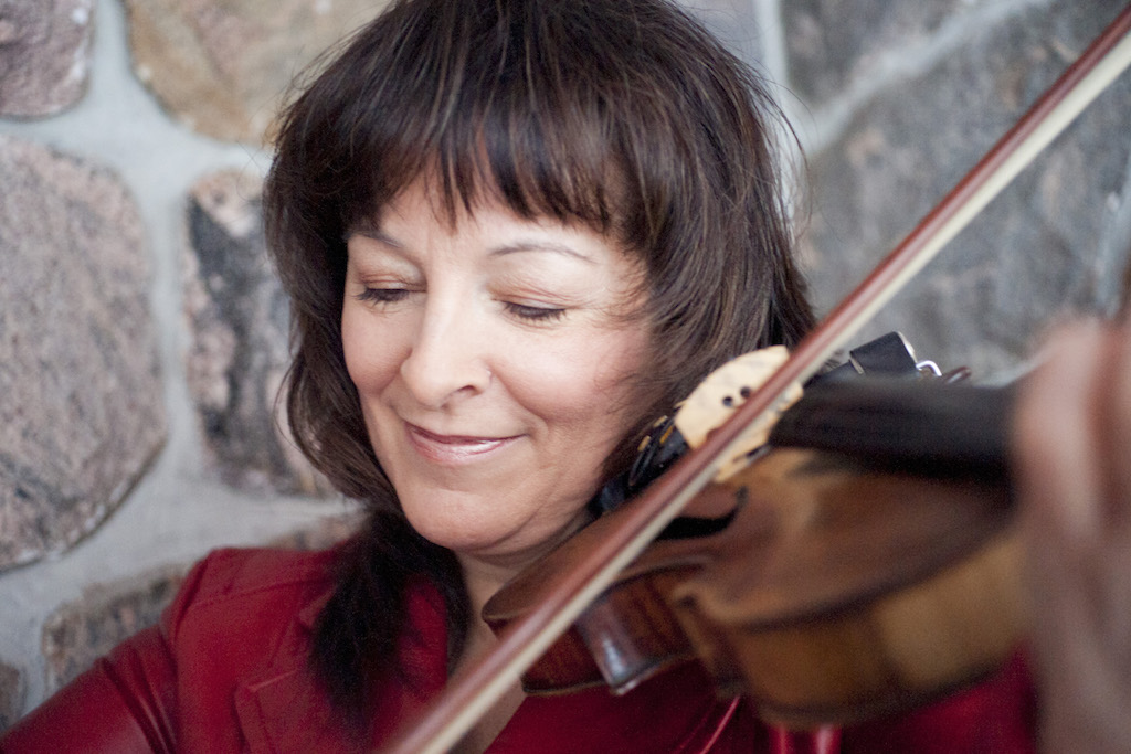 Cindy smiling and playing the fiddle in front of a stone wall.