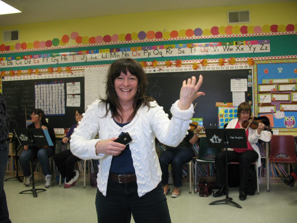 Cindy holding an imaginary fiddle, and smiling, standing in the middle of a classroom.